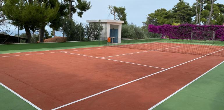 Campo da tennis in terra rossa circondato da alberi e fiori.